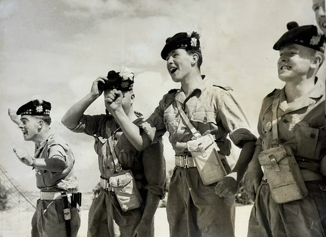 This is a photograph, not a designed graphic.

Soldiers in military uniform and Scottish caps laughing together outdoors, one holding a camera.