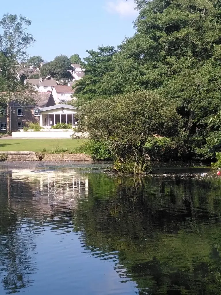 River view of a riverside building with a columned pavilion, surrounded by mature trees and hillside houses beyond.