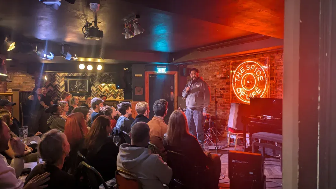 Comedian performing on stage to a packed crowd in an intimate comedy club with red neon signage and stage lighting.