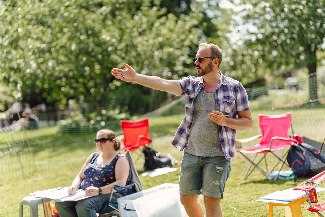 Man giving a presentation or leading a workshop outdoors in a sunny garden, with seated audience members and pink camping chairs visible.