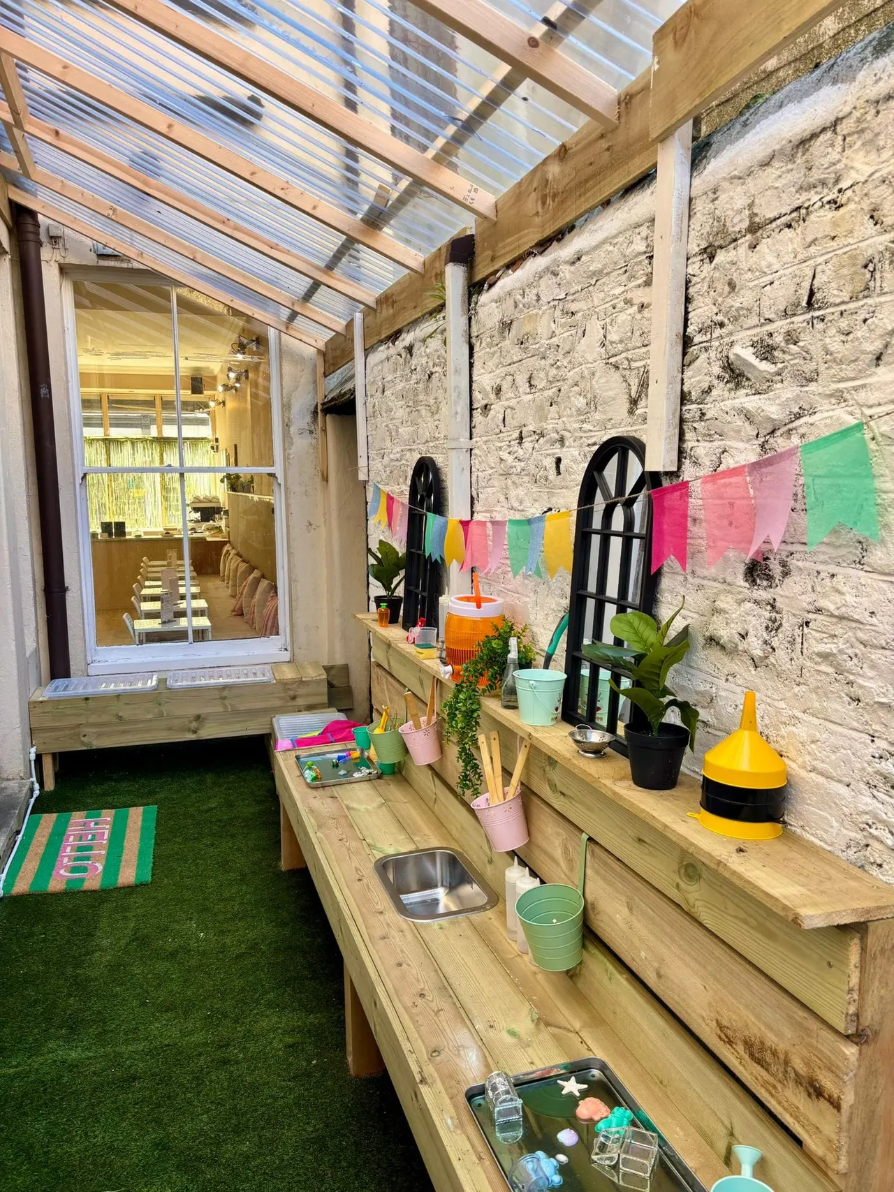 Children's outdoor play area with wooden mud kitchen, colourful bunting, potted plants and stone wall under polycarbonate roof.