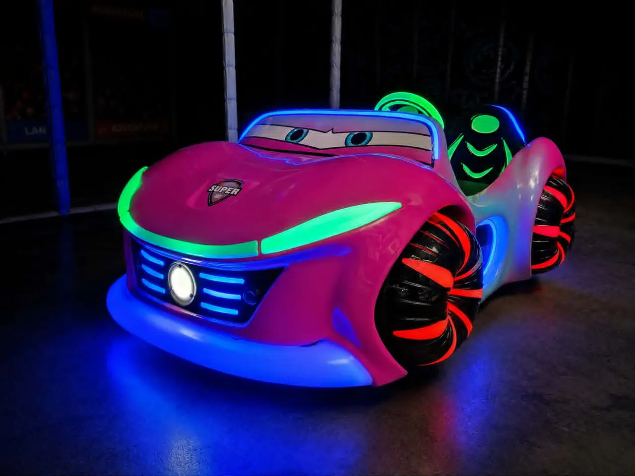 Brightly lit children's ride car in magenta with neon blue and green trim, featuring a character face design and oversized wheels.