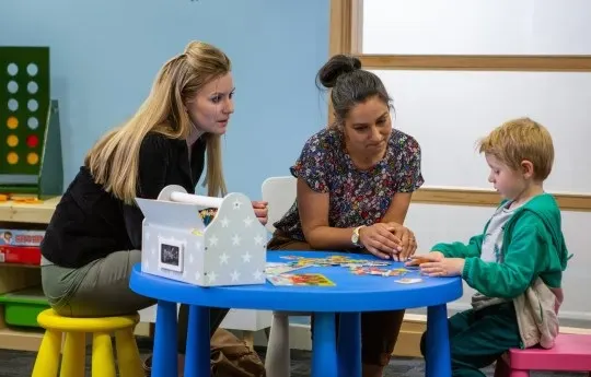 Two adults and a young child work together at a blue table during an indoor activity session.