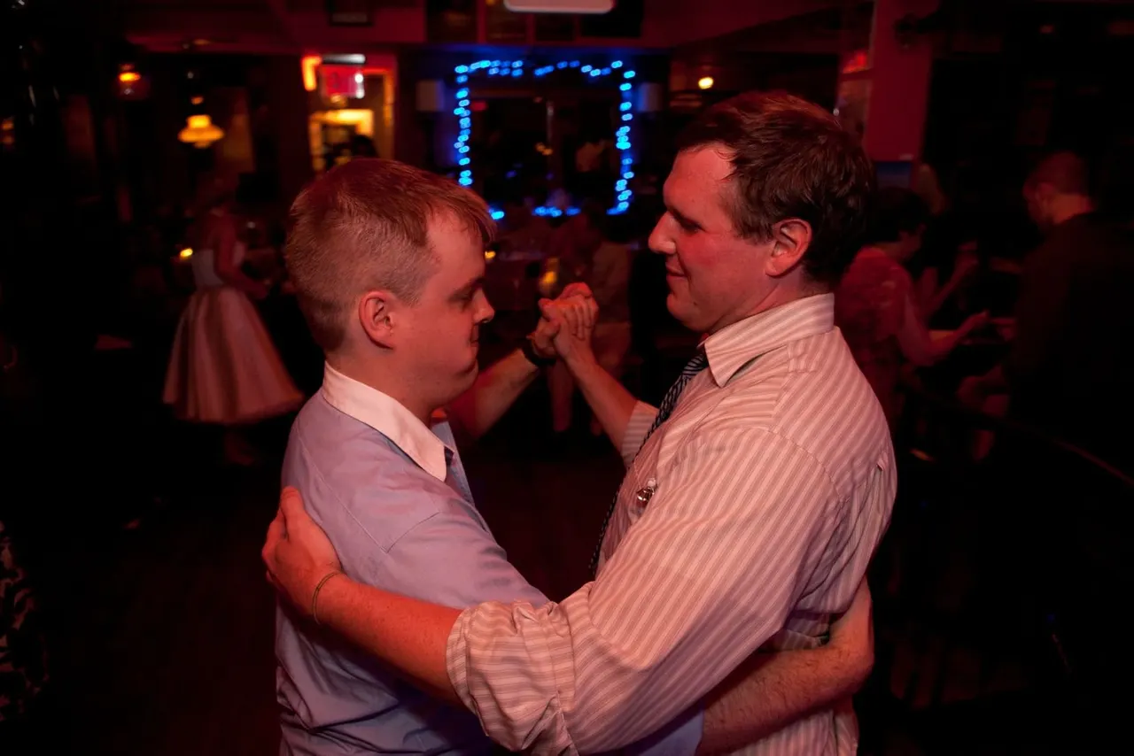 Two men dancing together in a dimly lit venue with red and blue neon lighting and other guests in the background.