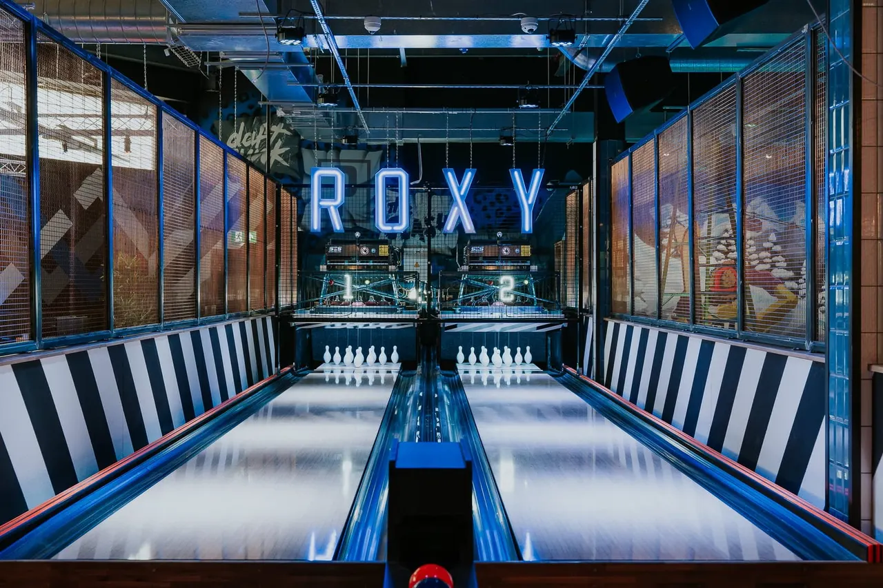 Two bowling lanes with white pins set up, blue and red trim, neon 'ROXY' sign, striped side panels, and arcade machines visible in the background.