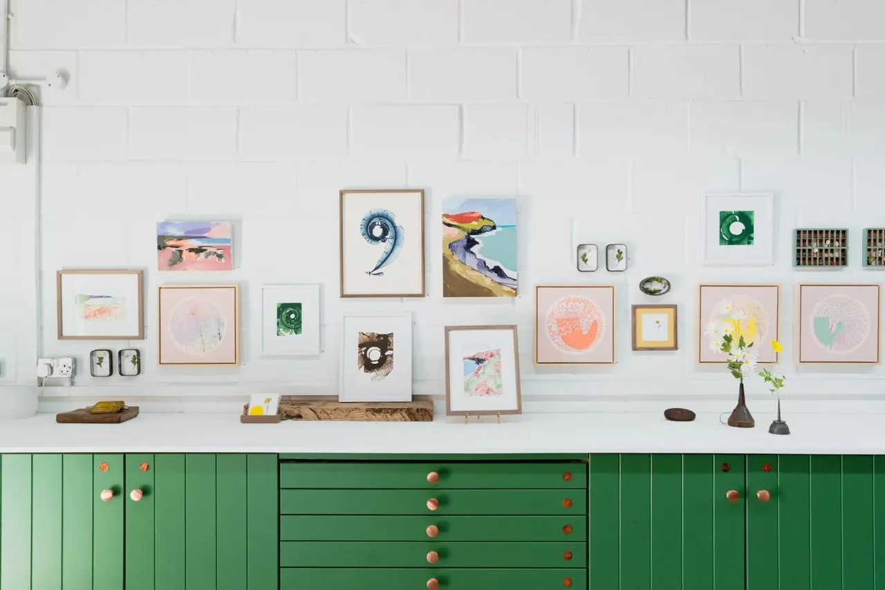 Display of framed artworks and decorative objects arranged on white wall above green cabinetry with wooden handles.