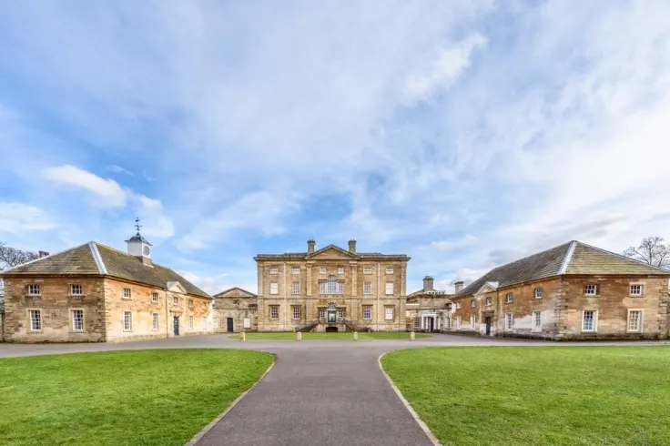 A grand stone mansion with symmetrical wings viewed across a manicured lawn and curved driveway under a partly cloudy sky.