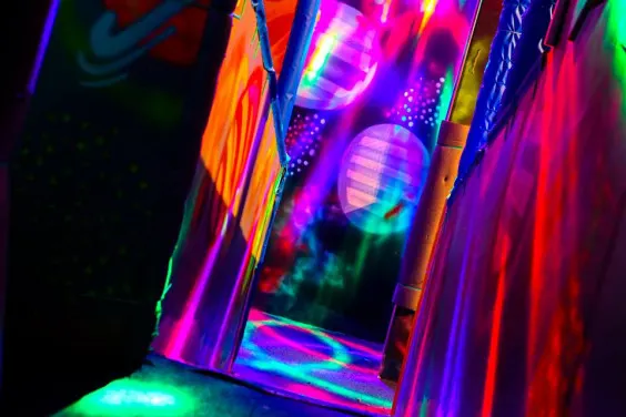 Person in neon glow-in-the-dark play environment with bright pink, purple, orange and green lights.