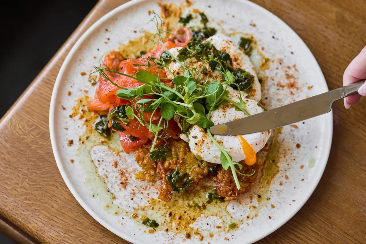 Plated dish with whipped cheese or cream base, topped with red peppers, green leaves, microgreens and spiced oil, being cut with a knife.