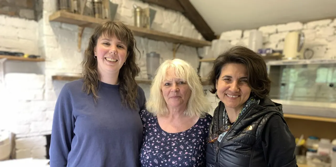 Three women smiling at the camera inside a historic pottery workshop with shelves and equipment.