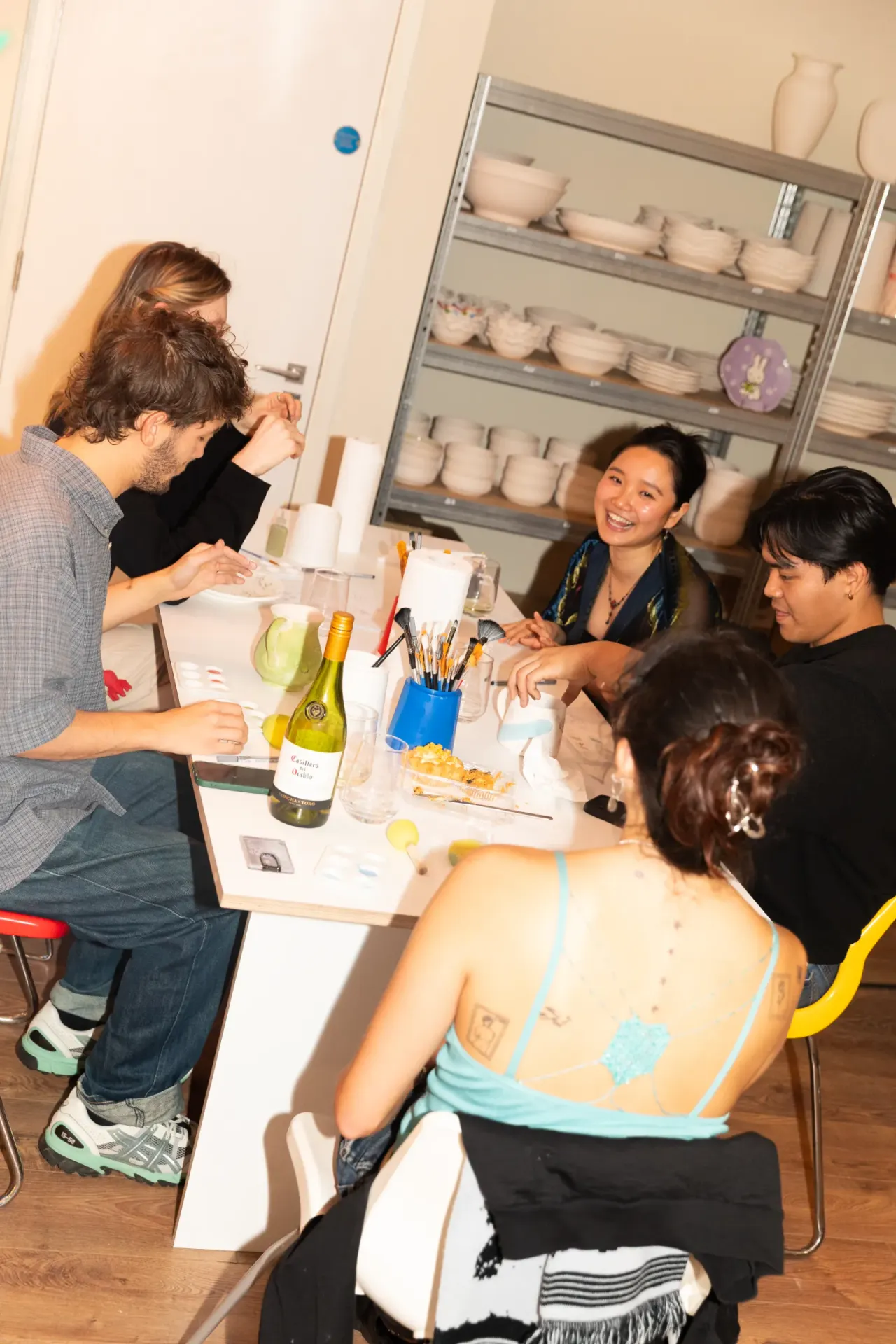 A group of four people sit around a table during a pottery painting workshop, with wine, paintbrushes and supplies in front of them.