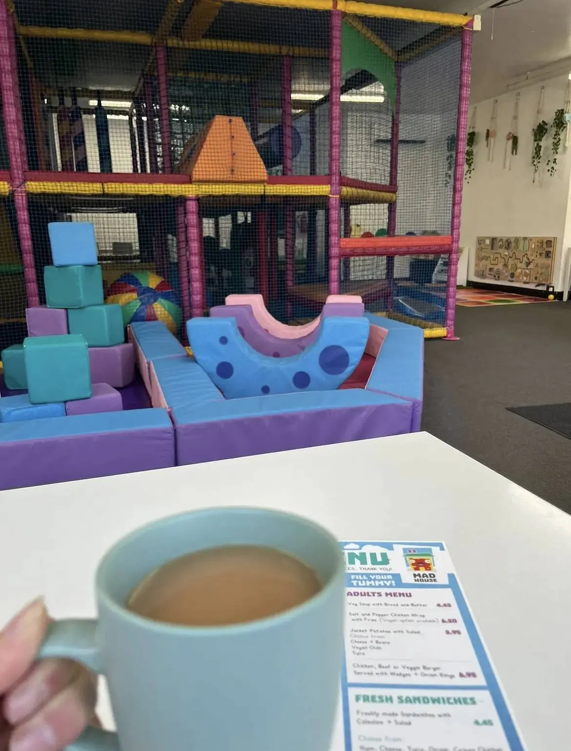 A hand holds a light blue mug of hot beverage in an indoor play area with colourful foam structures and a large mesh-enclosed play frame.