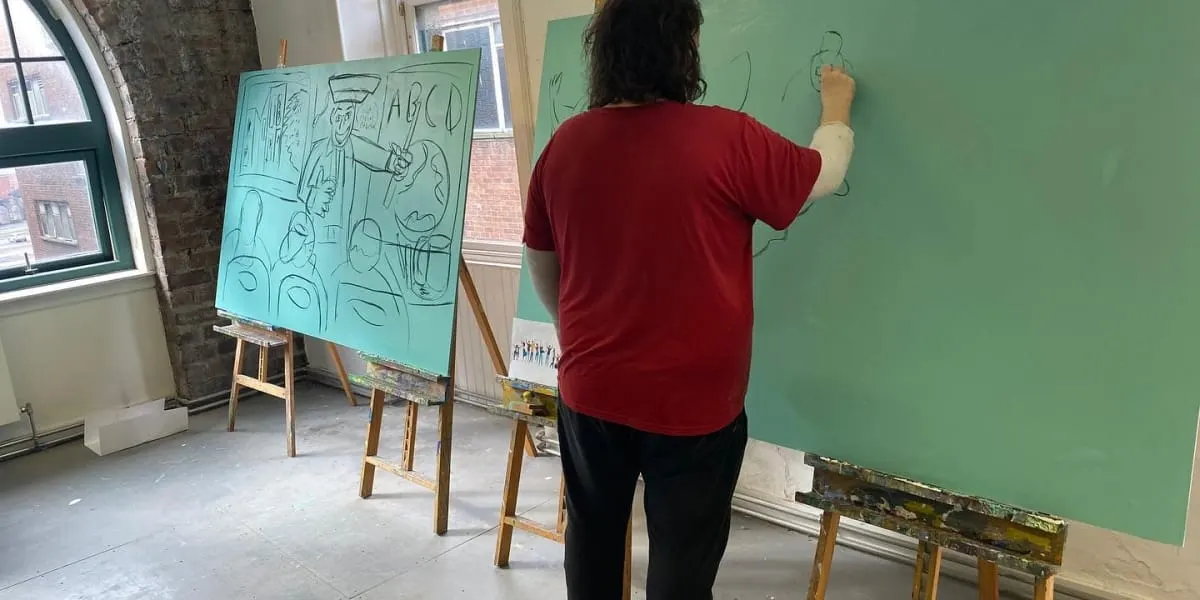 Person in red shirt drawing on green wall beside easel with sketch pad in art studio.