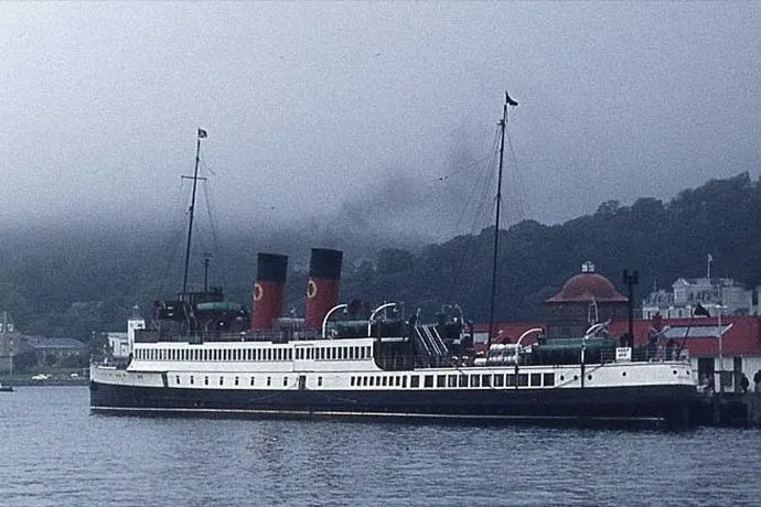 Historic paddle steamer with red funnels moored at a misty waterfront with forested hills beyond.