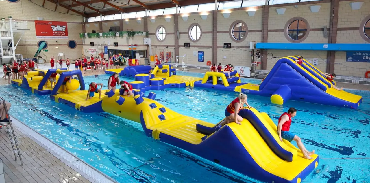Children in red swimming attire navigate inflatable obstacle course structures in an indoor swimming pool.