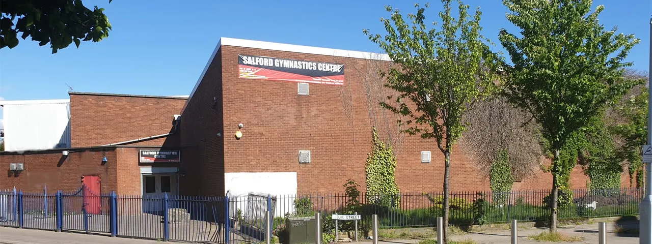 Brick building with "Salford Gymnastics Centre" signage, surrounded by blue fencing and green trees under clear sky.