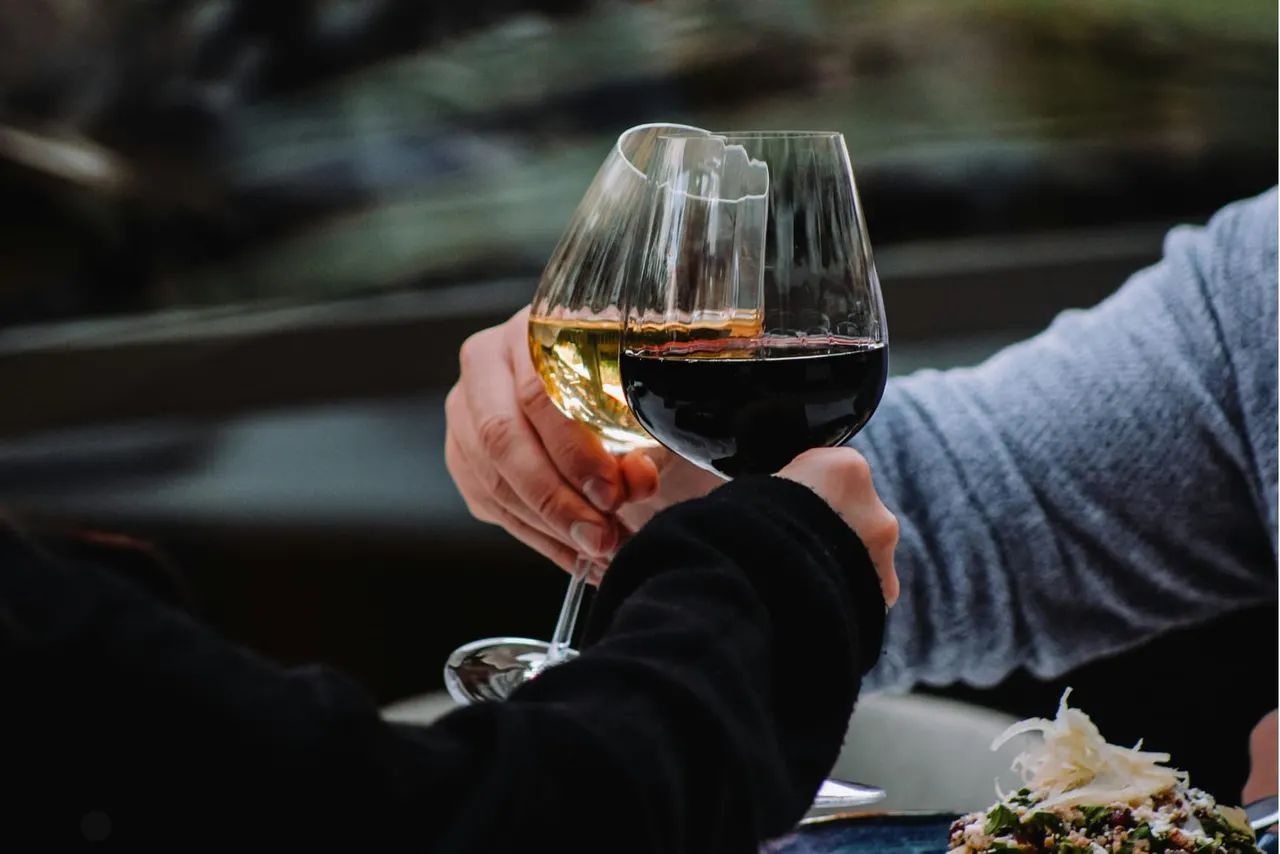 Person holding two wine glasses, one with white wine and one with red wine, outdoors with blurred woodland background.