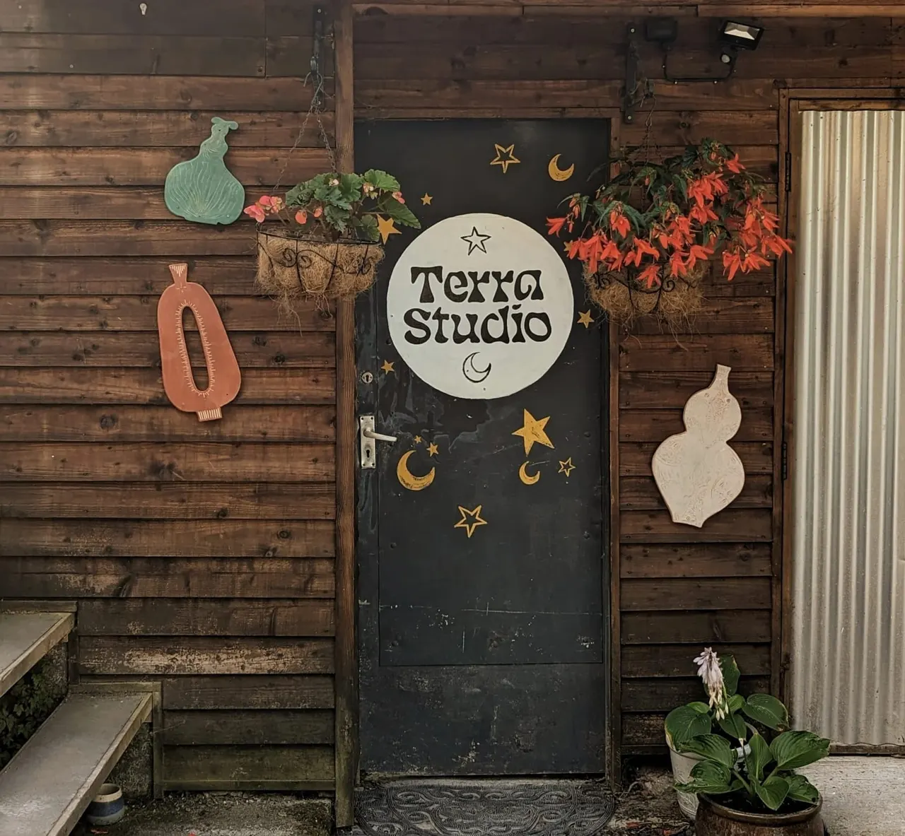 Entrance to Terra Studio with black door decorated with gold stars and moons, flanked by dark wooden walls adorned with hanging planters, ceramic vessel decorations, and autumn foliage.