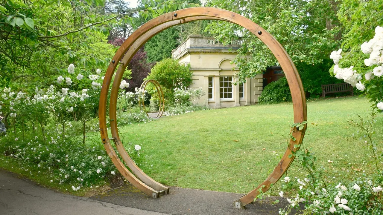 Wooden circular arch framing a garden lawn with white flowering shrubs, stone building and ivy-covered walls beyond.