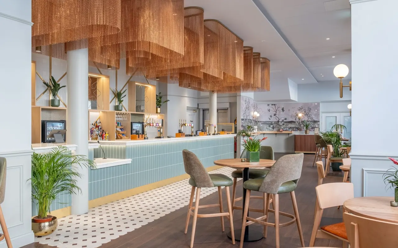 Modern hotel or leisure venue bar and lounge area with wooden furniture, plants, and a contemporary service counter.