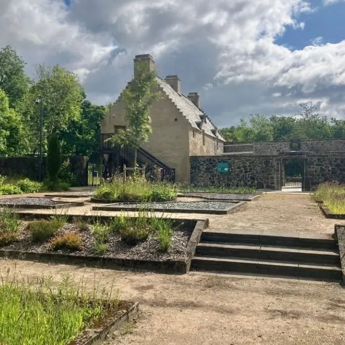 Historic stone cottage with modern landscaped grounds featuring raised beds and steps, surrounded by trees and stone walls.