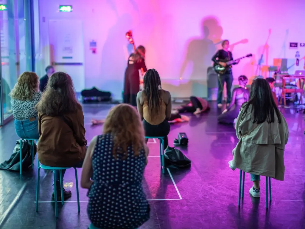 Audience members seated on stools watch a live band perform on an illuminated stage in purple lighting.