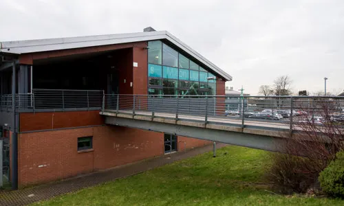 Modern brick building with large windows and metal railings beside a grassy area.