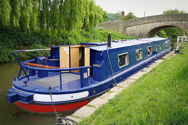 Blue and red narrowboat moored on a calm canal with stone bridge and willow trees in background.