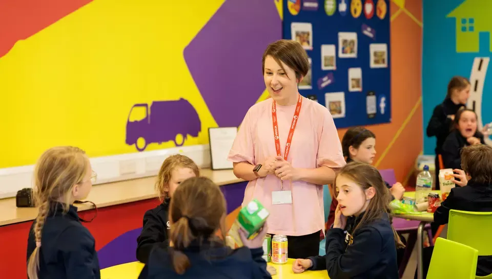 Staff member in pink shirt leads activity with children sat around colourful table in bright, playful indoor learning space.
