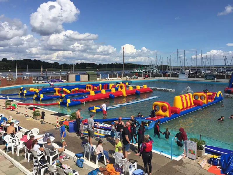 Children and adults enjoying inflatable obstacle courses and swimming in a waterfront leisure pool with moored sailing boats visible beyond.