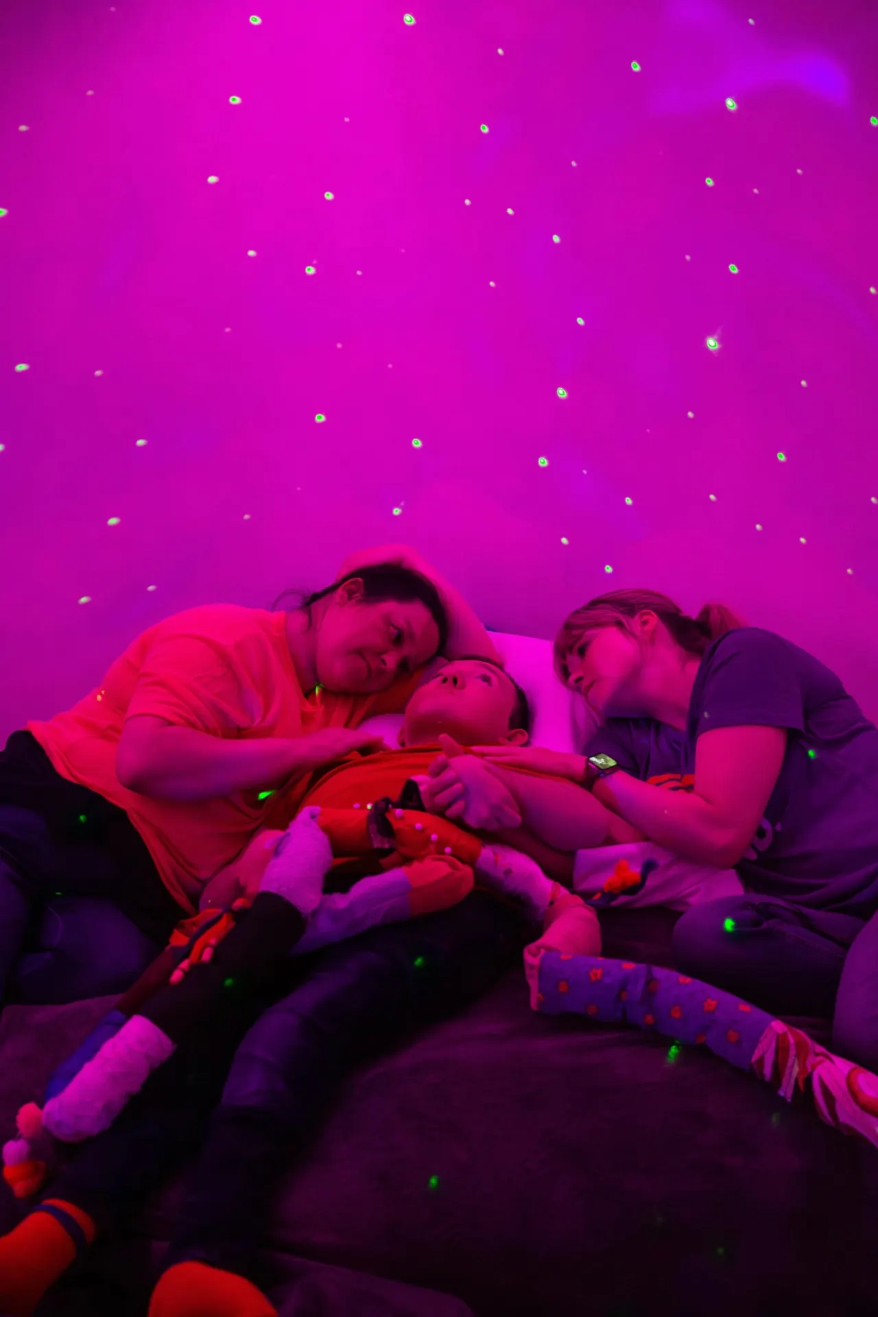 Three children lying on a padded surface under vibrant magenta and purple neon lighting with floating glowing particles.
