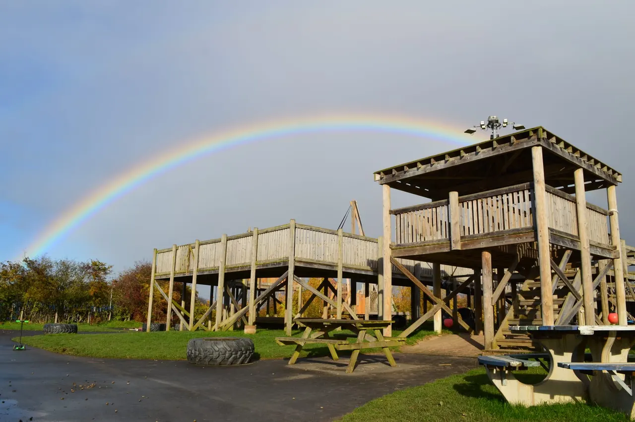 Wooden adventure playground structure with viewing tower beneath a rainbow, set in parkland with autumn trees.