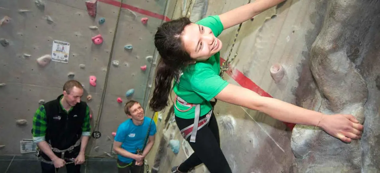 A young woman in a green shirt climbs an indoor rock wall whilst two instructors watch from below.