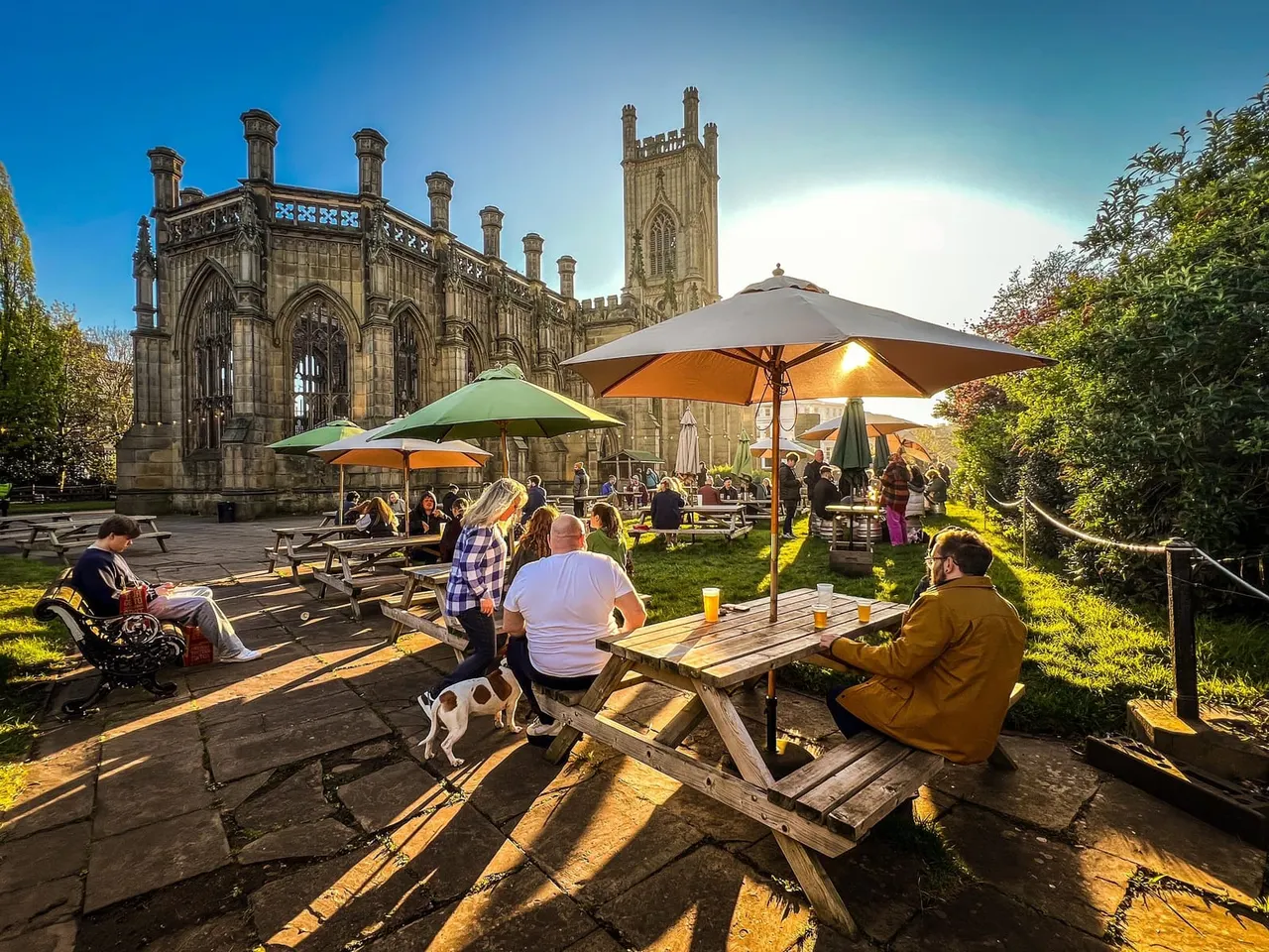 People enjoying drinks and conversation at picnic tables with coloured umbrellas in a garden, with a historic stone church building behind them.