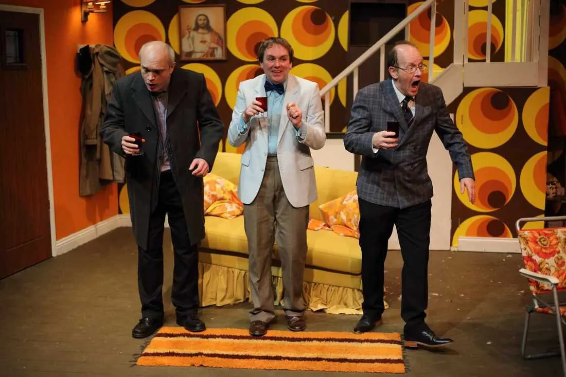 Three men in business suits perform a comedic scene on a theatre stage set decorated with orange and yellow circular designs.