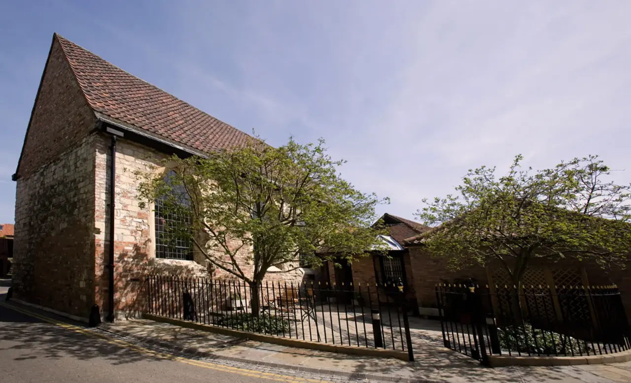 Historic stone building with pitched terracotta roof and iron railings, surrounded by trees on a sunny day.