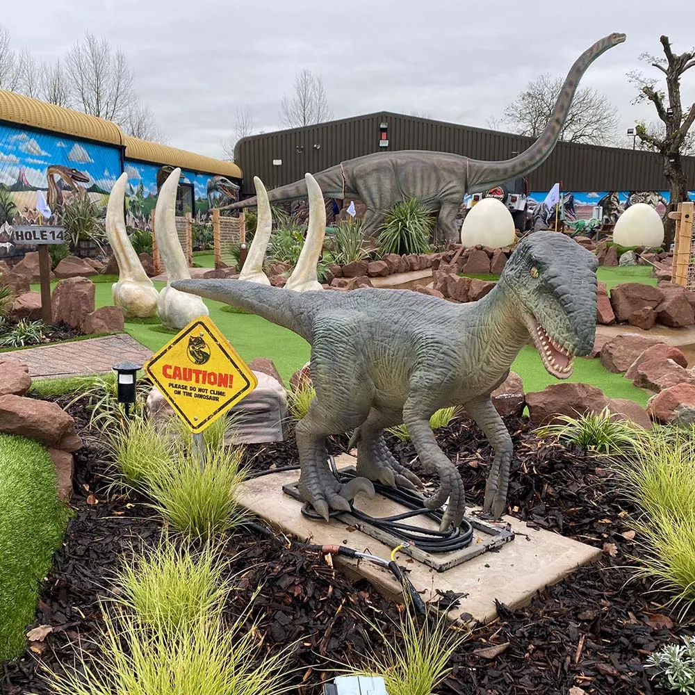 Dinosaur-themed mini golf course with large model dinosaurs, eggs, tusks and a caution sign among artificial plants and landscaping.