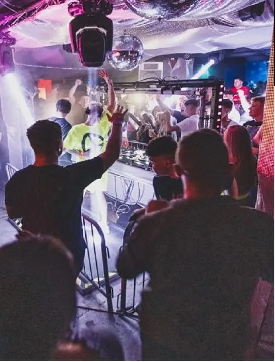 Crowded nightclub interior with people dancing under colourful purple and pink lighting with a DJ booth.