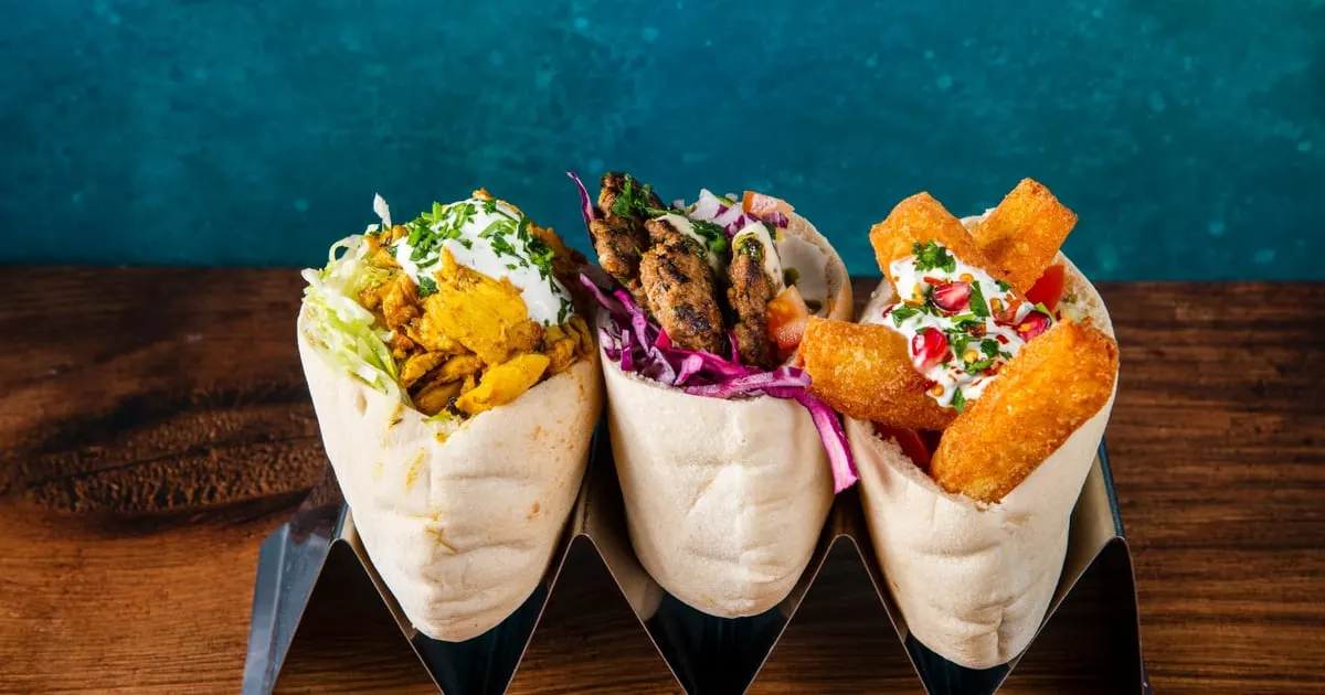 Three filled wraps with various toppings including spiced vegetables, meat, and fried items arranged on a wooden table against a teal wall.