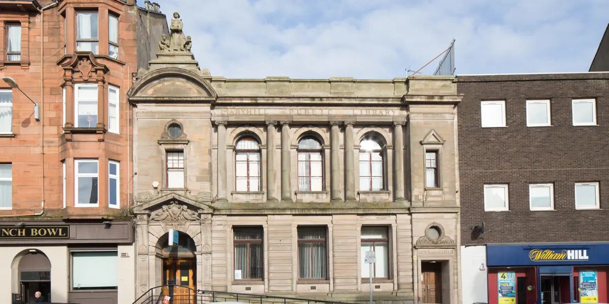 A street view of historic stone buildings including a sandstone library with arched windows flanked by red tenement and brick structures.