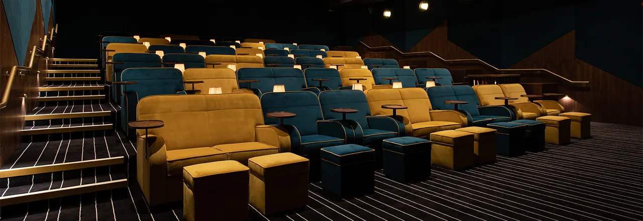 Empty cinema auditorium with rows of blue and gold reclining seats and footrests lit by warm overhead lighting.