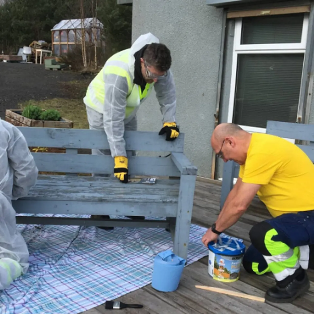 Workers in high-visibility clothing painting a wooden bench outdoors on a patio area.