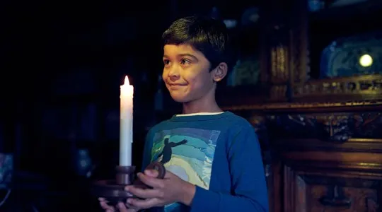 Young child holding a lit candle indoors at an atmospheric historic house setting.