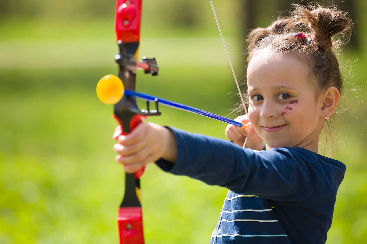 Young child with face paint aiming a toy bow and arrow at a yellow ball target outdoors on grass.
