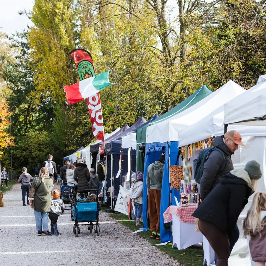 Visitors browse market stalls and tents at an outdoor autumn festival surrounded by trees.