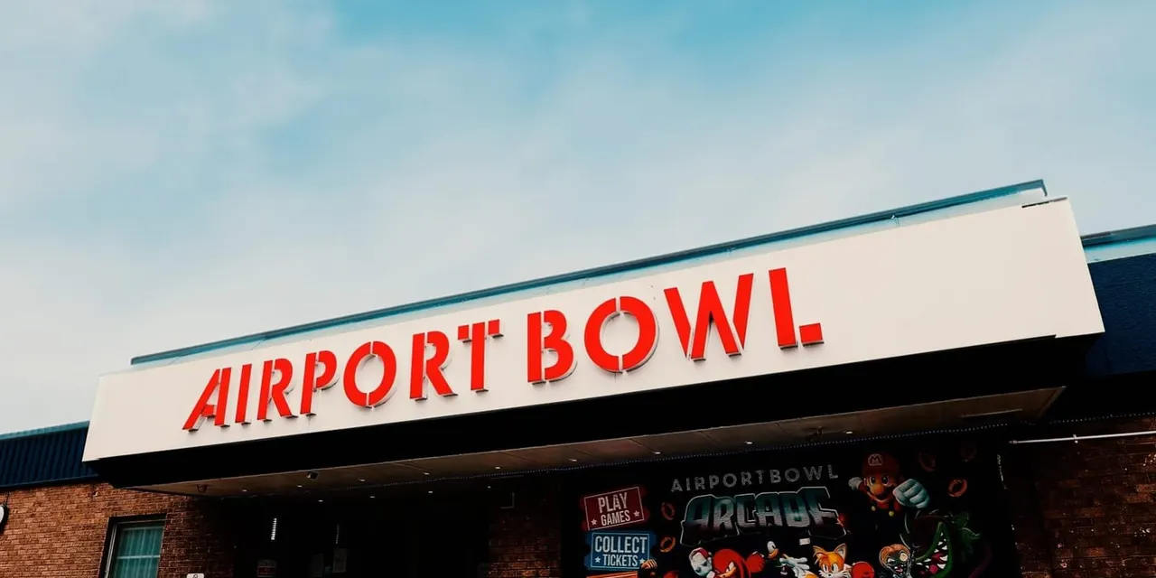 Red and white "AIRPORT BOWL" signage on a building exterior with blue sky above and brick walls below.