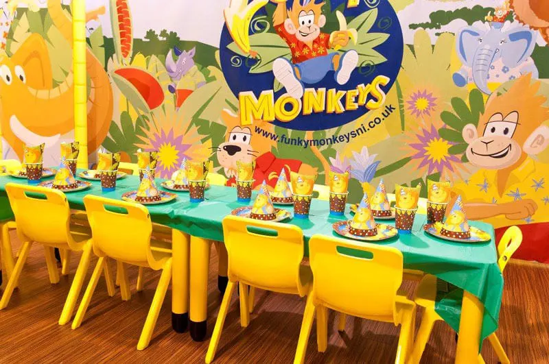 A children's party room with a long table set with yellow chairs, colourful place settings, and a jungle-themed backdrop featuring cartoon monkeys and animals.