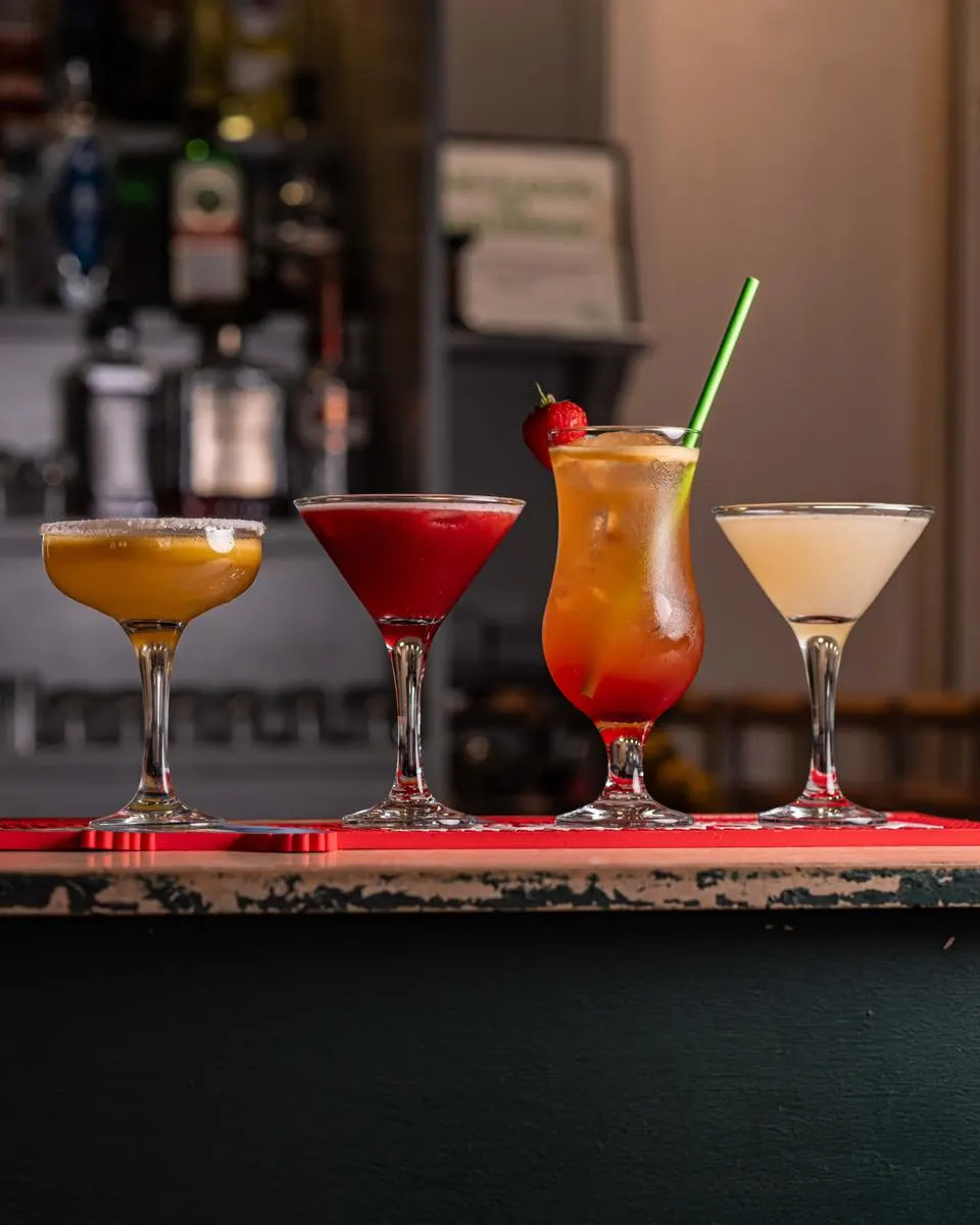 Four colourful cocktails displayed on a bar counter with bottles blurred in the background.