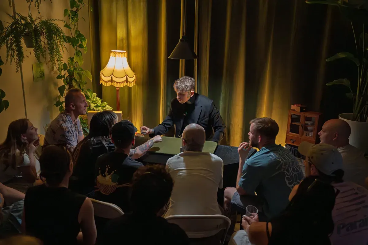 A magician performs an intimate close-up magic trick for a seated audience of about a dozen people in a warmly lit room with vintage décor and potted plants.
