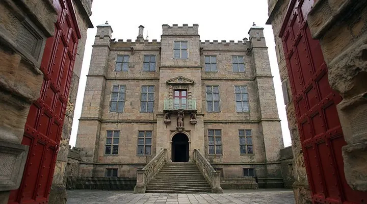 Historic stone castle with crenellated towers and multiple windows, viewed through large red gates from the courtyard.